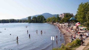 Una playa junto al lago con gente nadando y tomando el sol, un pabellón y edificios al fondo.