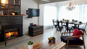 A modern living room with a fireplace, a dining area with a chandelier, and a flat-screen TV mounted on the wall. - A favorite chalet in Orford (Orford)