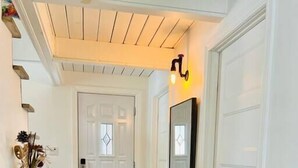 A hallway with a wooden ceiling, a white door, a wall-mounted light, and a small table with a vase and papers. - A favorite chalet in Orford (Orford)