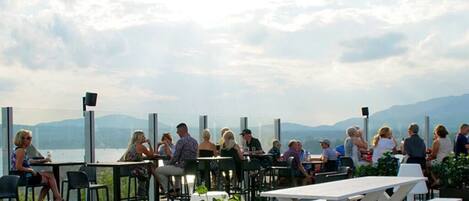 An outdoor dining area with tables and chairs, overlooking a body of water and mountains.