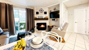 A living room with a fireplace, a dining table with fruit, a sofa, and a TV mounted on the wall. - Merry Sud  Suite-Condo with Indoor Pool and Spa (Magog)