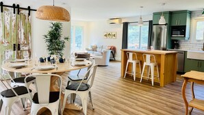 Eine geräumige Küche mit Holzboden, einer zentralen Kochinsel mit Barhocker und einem Essbereich mit weißen Stühlen und einem runden Tisch.