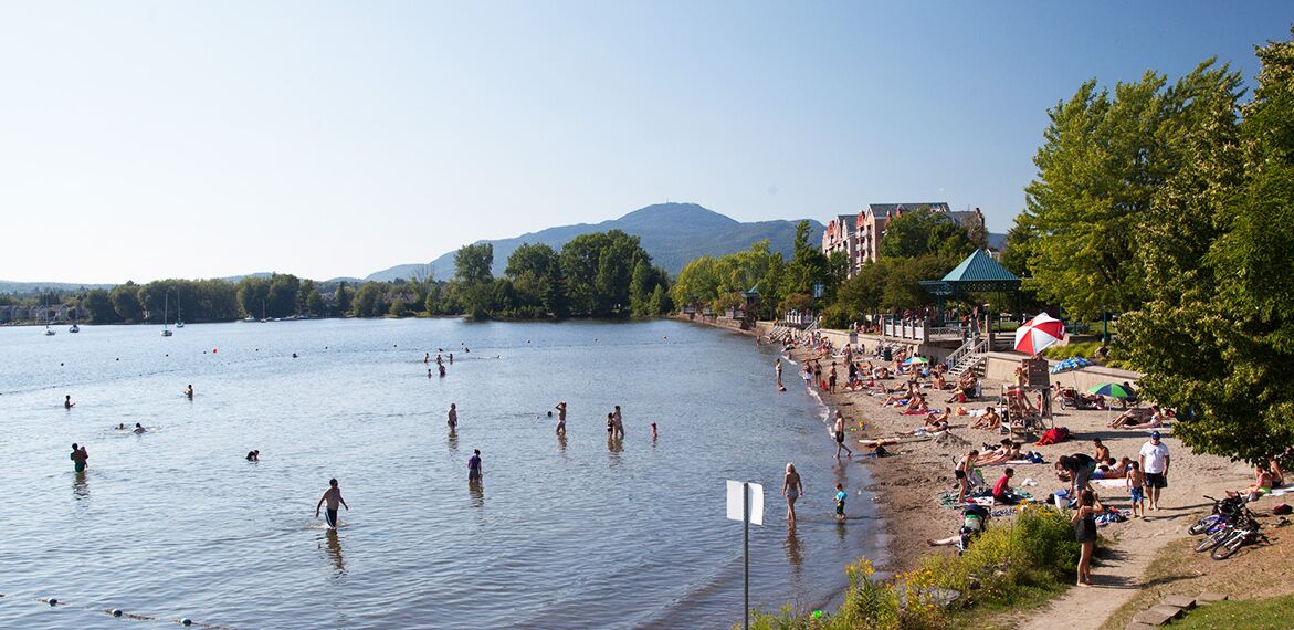 A beach with people swimming and sunbathing, a large umbrella, and a building with a green roof in the background.