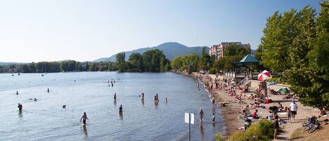 A beach with people swimming and sunbathing, a large umbrella, and a building with a green roof in the background.