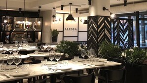 Interior de restaurante com mesas preparadas para refeições, área de bar com garrafas e parede decorativa com o nome 'ALESSA'.