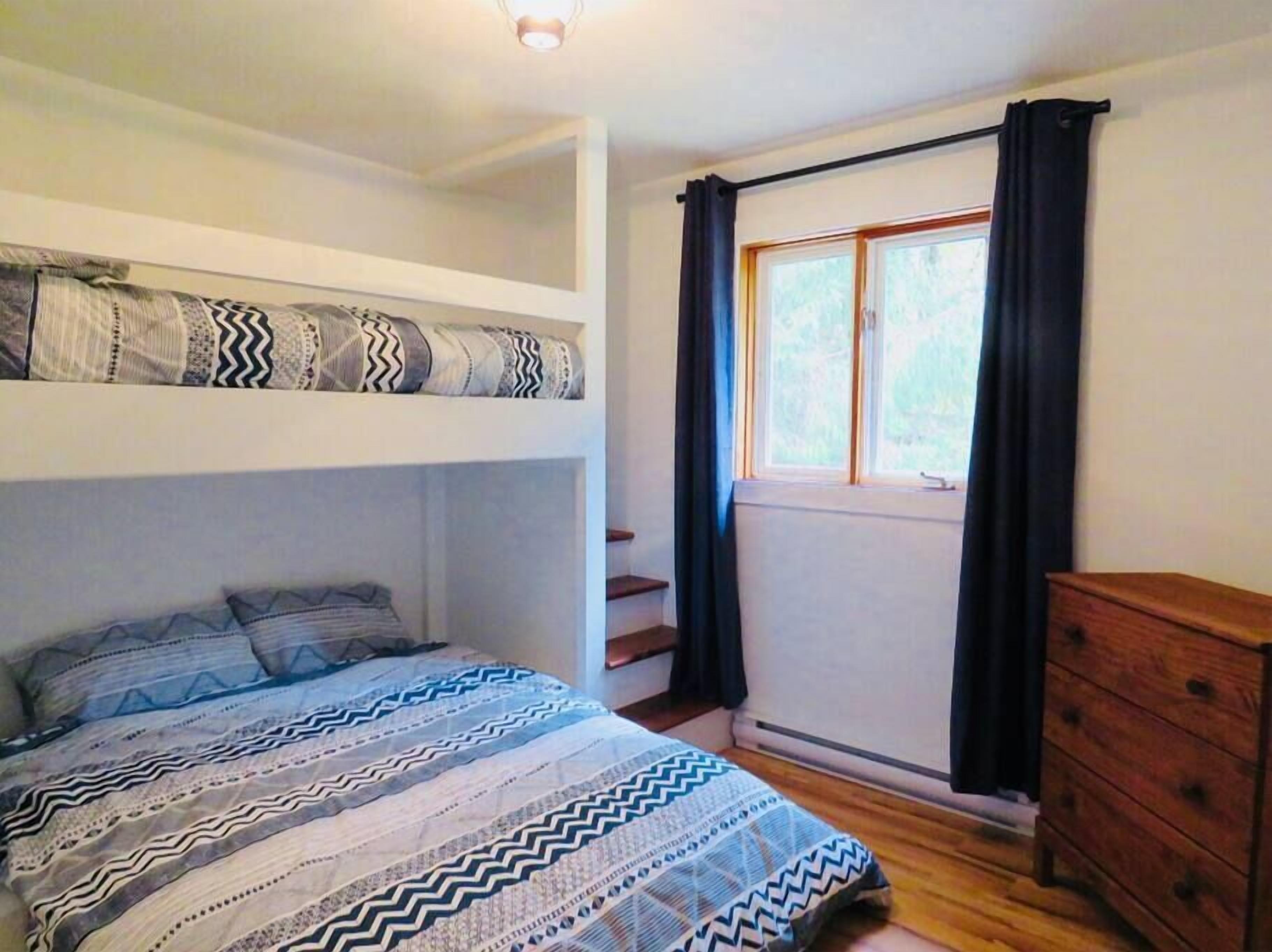 Ein Schlafzimmer mit Etagenbett, einem Einzelbett mit gemusterten Betttextilien, einer Holzkommode und einem Fenster mit Vorhängen.