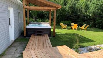Une terrasse en bois menant à un jacuzzi situé sous une pergola, avec des chaises d’extérieur et une pelouse.