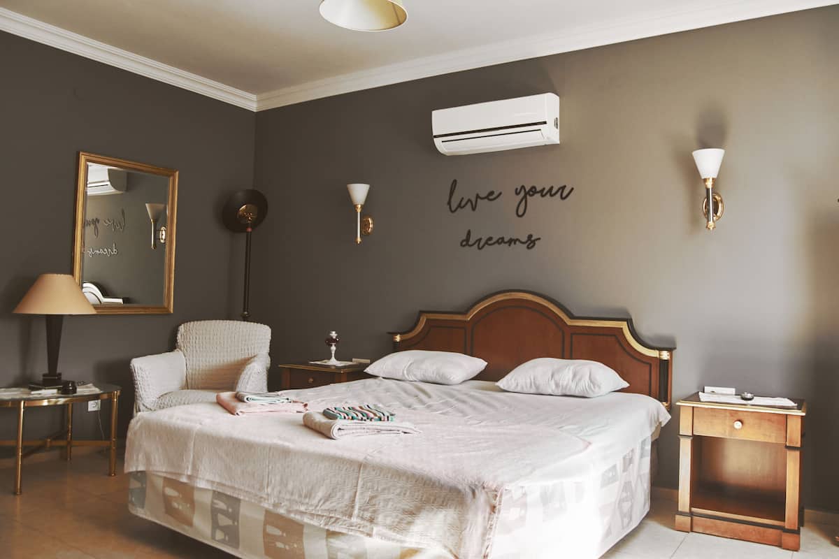 Een keurig opgemaakt slaapkamer met een bed, nachtkastjes, een stoel en een luchtverkoeler aan de muur.