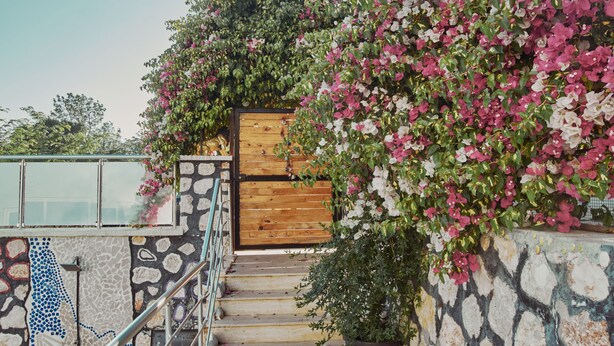 A staircase with a wooden railing, a stone wall with a mosaic pattern, and a wooden door surrounded by blooming pink and white flowers. - Villa Nordic Dream (Kas)