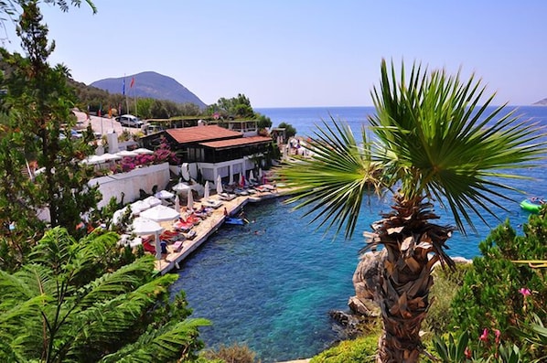 A coastal hotel with a poolside area, a mountain in the background, and a clear view of the sea.