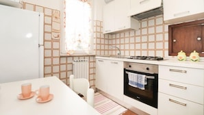 Een kleine keuken met witte kastjes, een gasfornuis, een ingebouwde oven, een raam met gordijnen met een patroon, en een radiator.