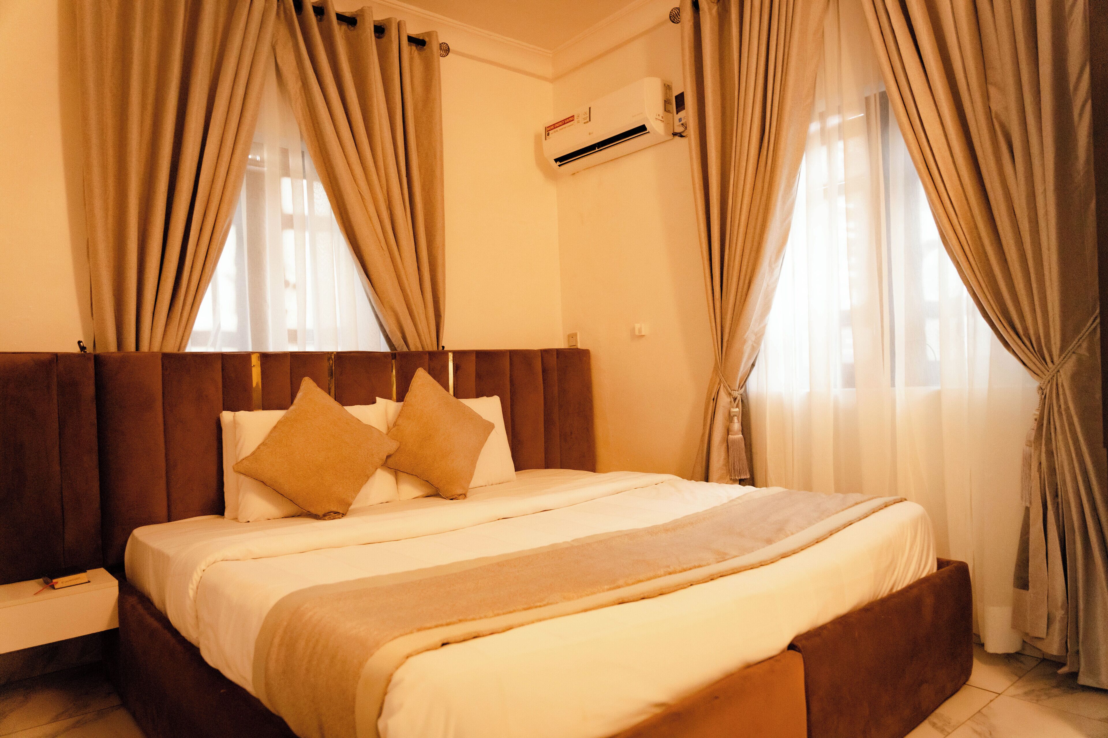 A hotel room with a large bed, beige curtains, an air conditioning unit, and a window with curtains.