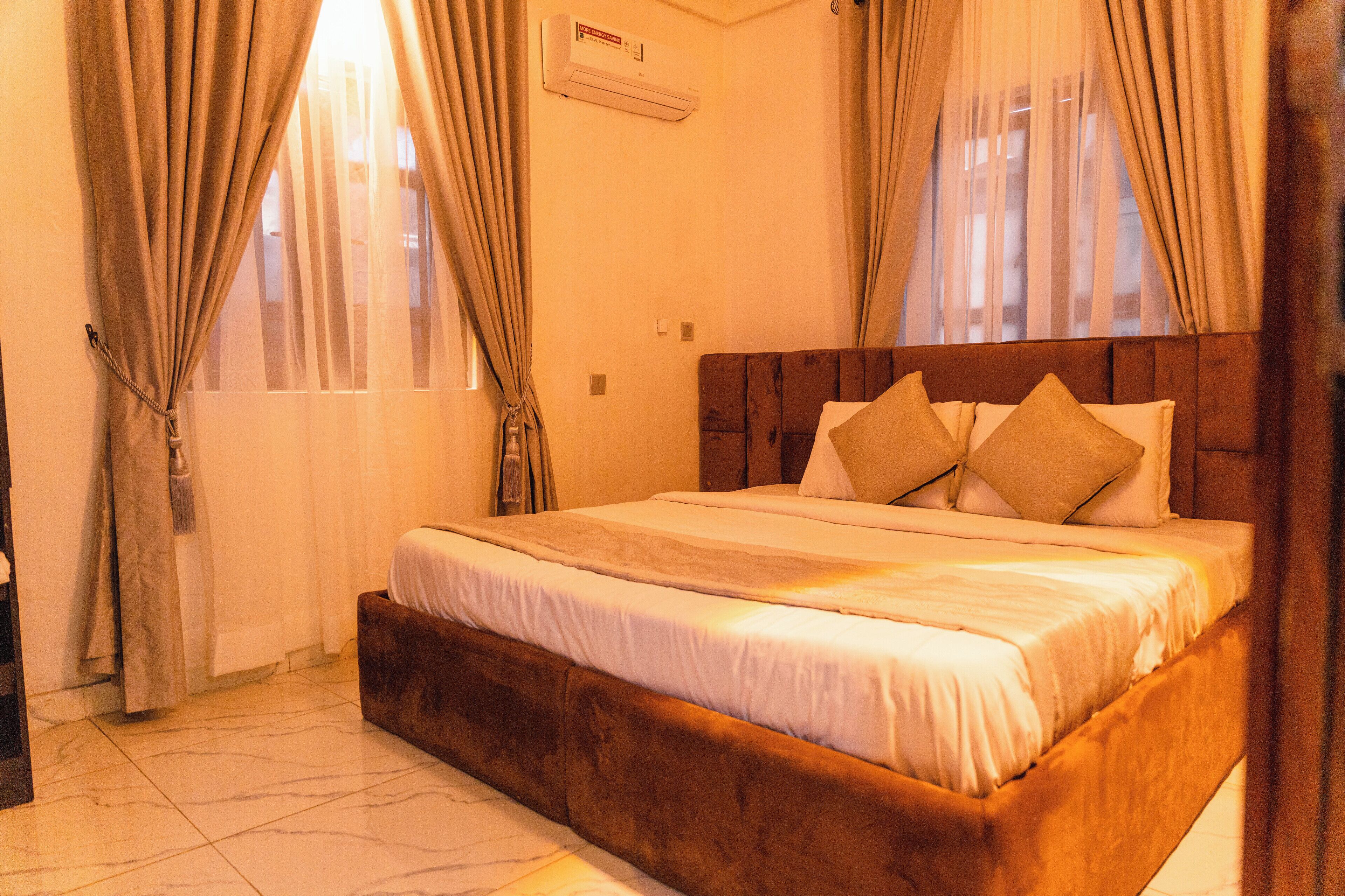 Uma cama bem arrumada com cabeceira de madeira, uma janela grande com cortinas e piso de azulejos.