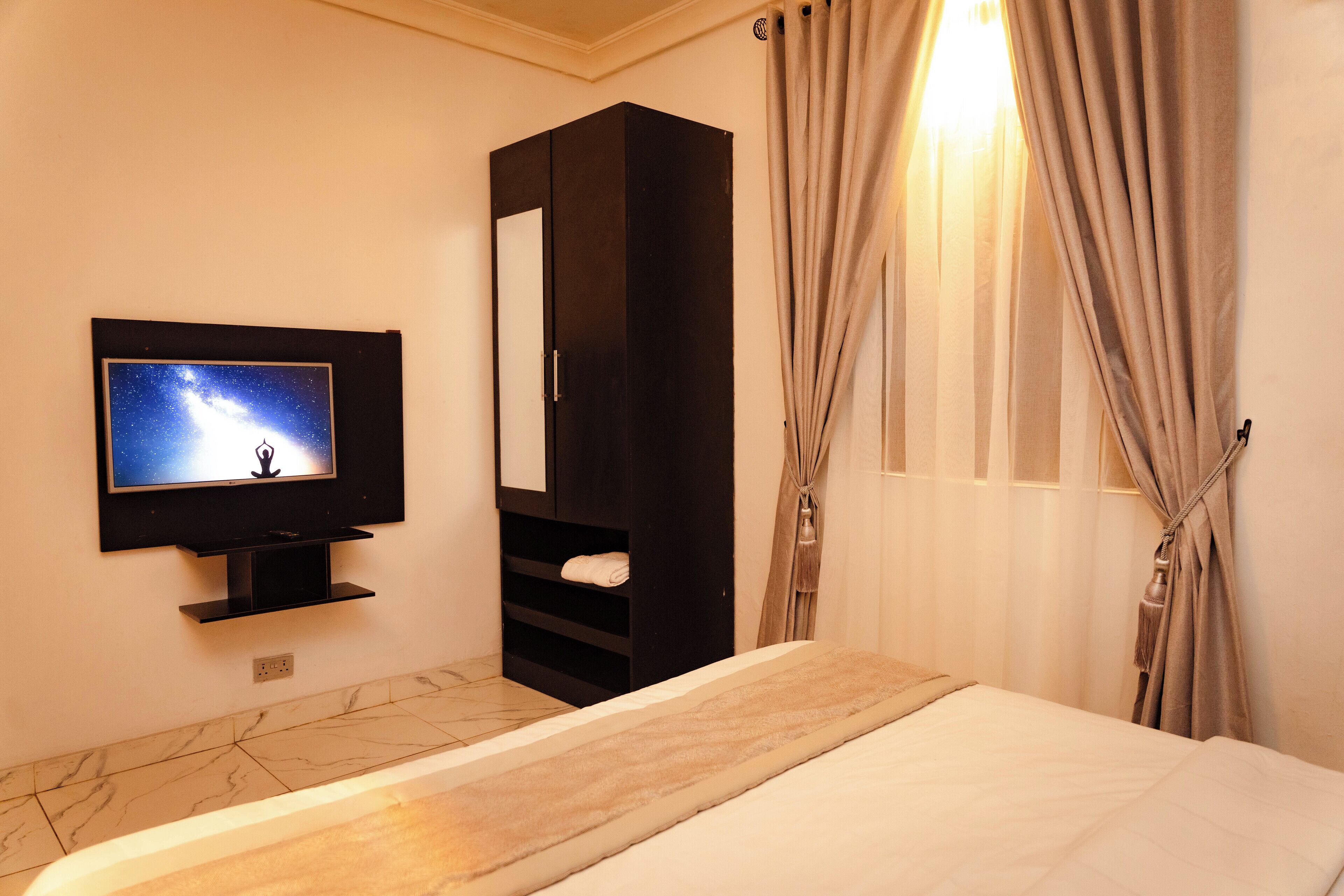 A hotel room with a bed, a TV mounted on the wall, a wardrobe, and a window with curtains.