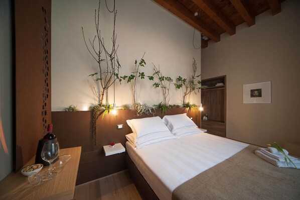 Cama bem arrumada com lençóis brancos, mesa de cabeceira com uma taça de vinho e um prato de biscoitos, e um arranjo de plantas fixado na parede.