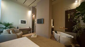 A modern hotel room with a bed, a sofa, a bathtub, a TV, and a large plant.