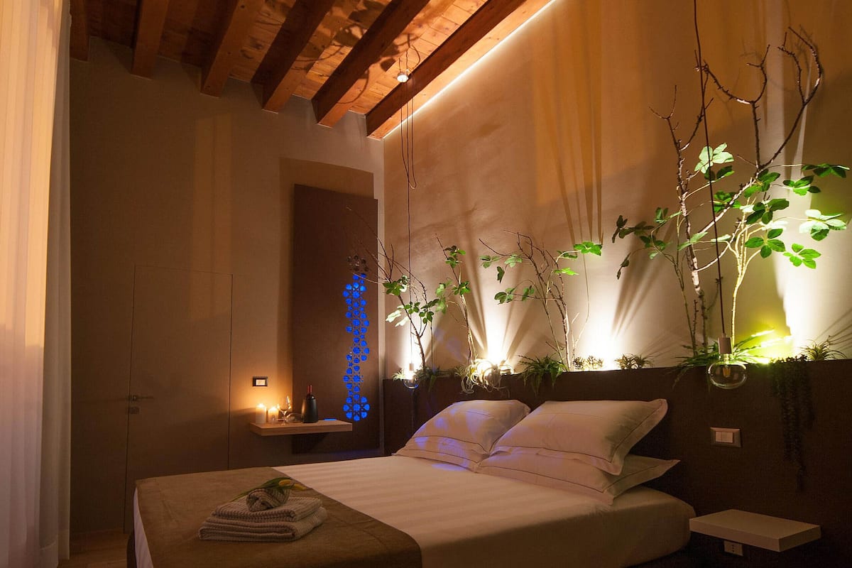 A bedroom with a wooden ceiling, a bed with white linens, a bedside table, and a wall-mounted plant.