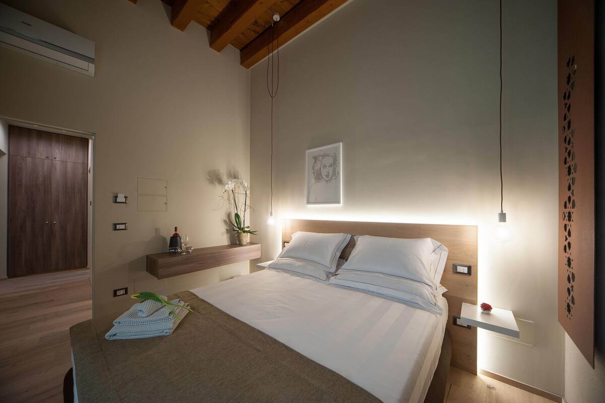 A bedroom with a wooden ceiling, a bed with white linens, a bedside table with a vase of flowers, and a wall-mounted shelf.