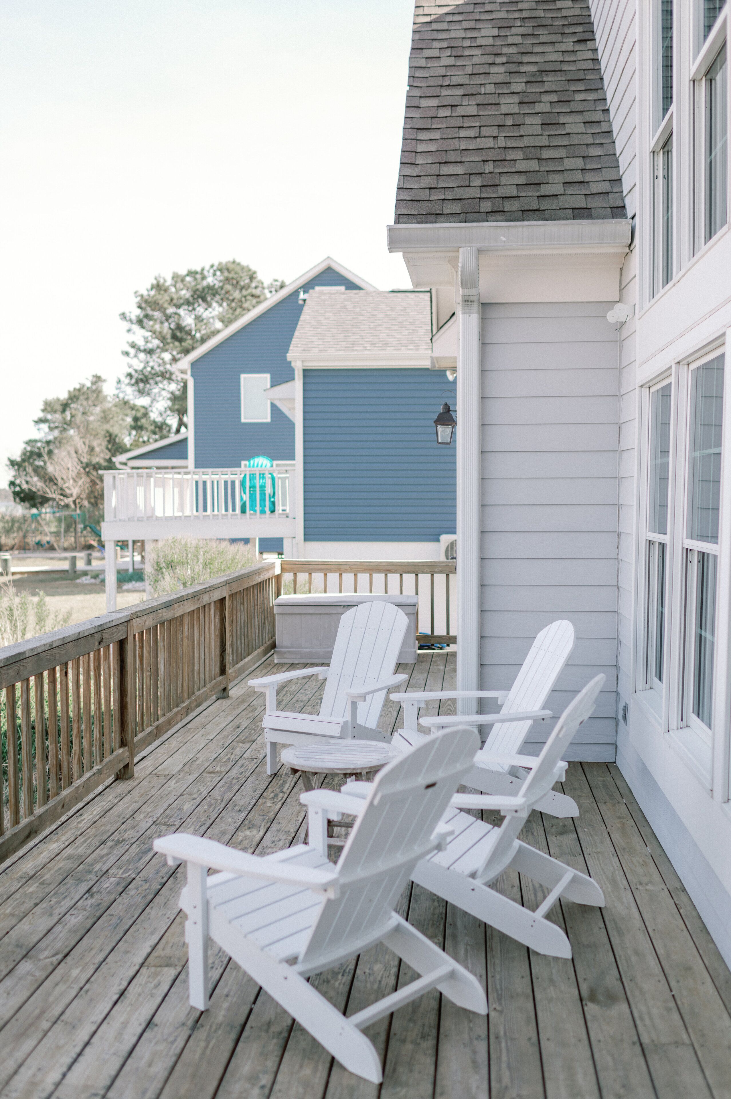 A wooden deck with white Adirondack chairs and a view of a blue house with a balcony.