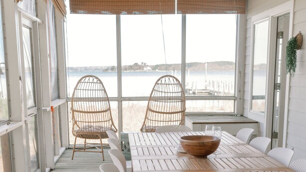 Une terrasse en bois avec une table et des chaises pour les repas, de grandes fenêtres et des fauteuils en osier.