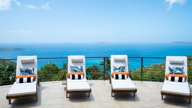 Una terraza con cuatro tumbonas, cada una con una almohada y un cojín, con vistas a la costa, el mar y unas islas distantes.