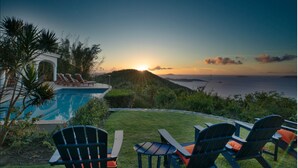Una piscina con sillas de descanso, una mesa y vistas al océano al atardecer.