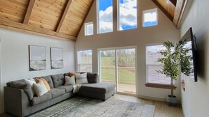 A spacious living room with a high ceiling, wooden beams, a gray sectional sofa, a large rug, and a potted plant.