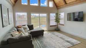 A spacious living room with a high ceiling, large windows, a ceiling fan, a sectional sofa, a flat-screen TV, and a potted plant.