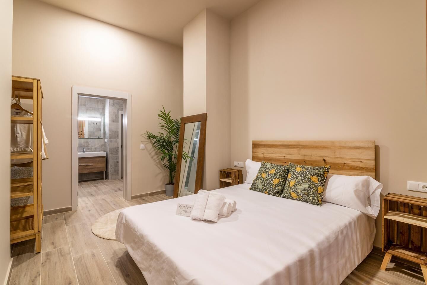 Une chambre avec une tête de lit en bois, un lit avec une literie blanche, une table de chevet, une armoire en bois et une salle de bain avec un miroir et un lavabo.