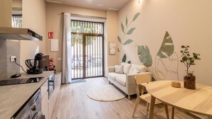 Una cocina moderna equipada con horno, cafetera y lavadora. Cuenta con una zona de comedor con mesa y sillas, un sofá y una planta decorativa.