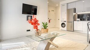 A modern kitchen with stainless steel appliances, a glass-top dining table with a vase of flowers, and a flat-screen TV mounted on the wall.
