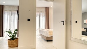 A modern hotel room with a large bed, a potted plant, and a view through a door into another room.