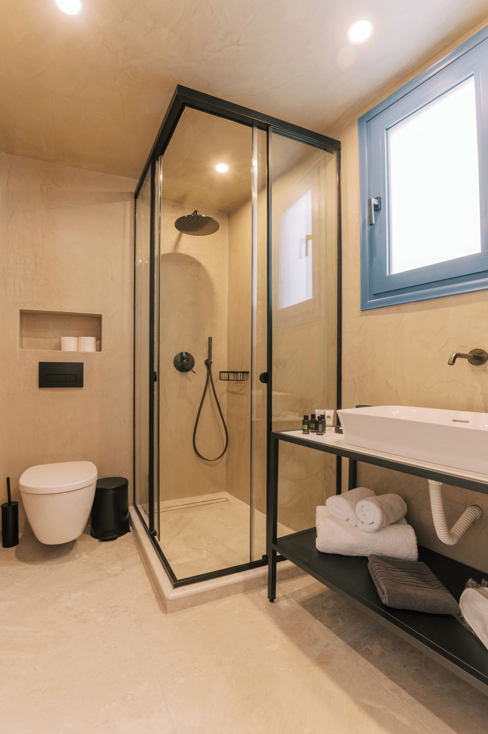 Een moderne badkamer met een douchecabine van glas, een wit toilet, een wastafel met toiletartikelen en handdoeken op een plank.