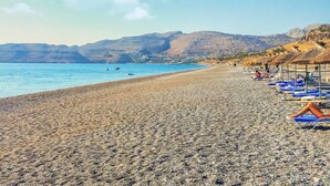 Una playa de guijarros con sombrillas y sillones de playa, un mar azul cristalino y colinas al fondo.
