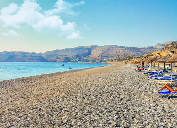 A pebble beach with umbrellas and lounge chairs, a clear blue sea, and hills in the background. - Kalathos Square Luxury Suites (Rhodes)