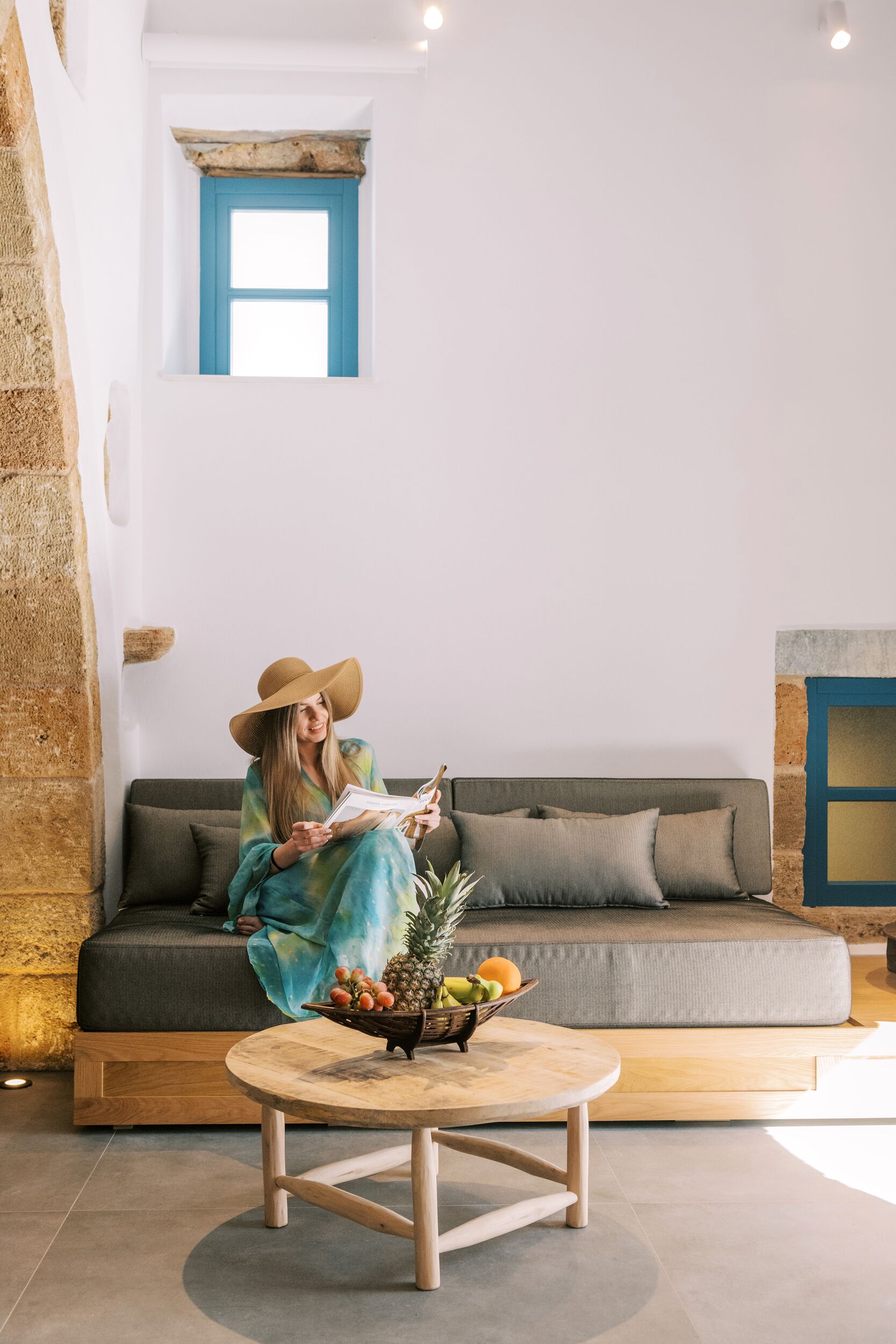 A person sitting on a grey sofa in a room with a stone wall, a small window with blue trim, and a wooden coffee table with a fruit bowl.