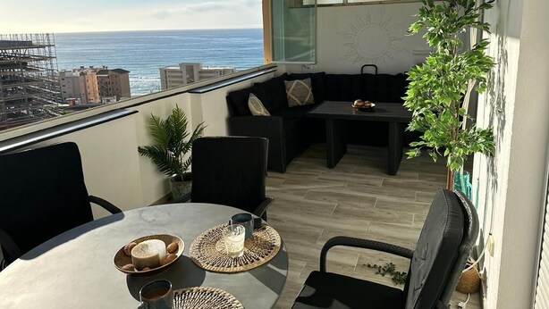 Un balcón con vista al océano, una mesa de comedor con platos y vasos, y un sofá negro.