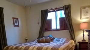 Une chambre à coucher comprenant un lit, un ventilateur de plafond, une fenêtre avec des rideaux, une table de chevet et une lampe.