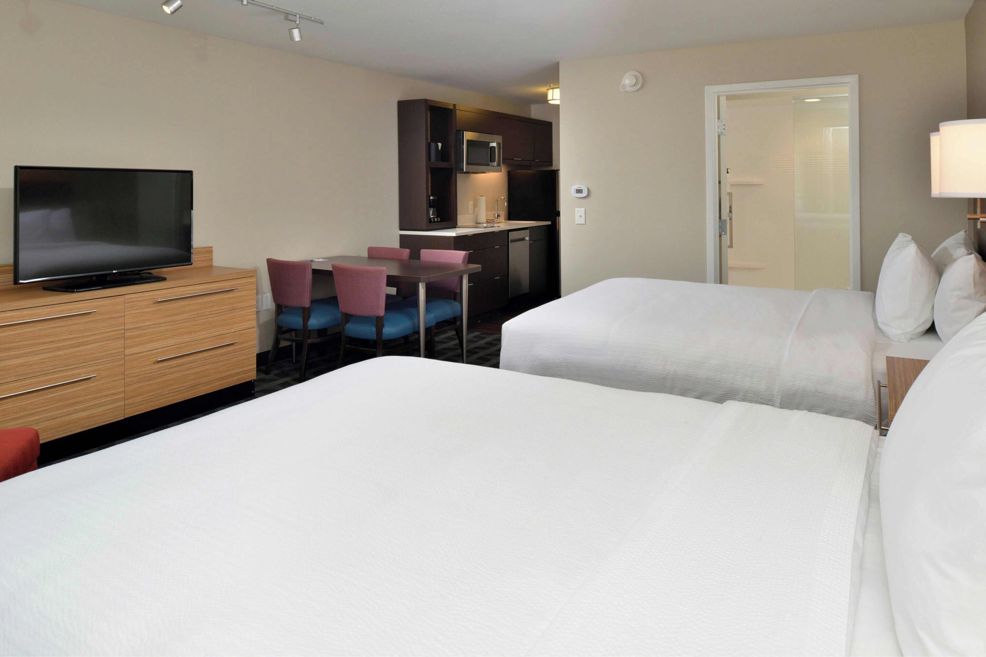 A hotel room with two beds, a TV, a small kitchenette, and a dining area.