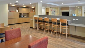 A modern dining area with wooden tables and chairs, a kitchen in the background, and a counter with stools.