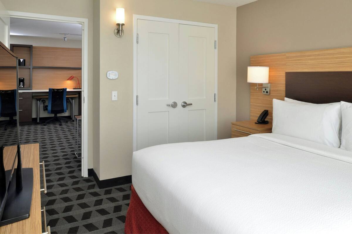 Une chambre d’hôtel avec un lit, un bureau et une chaise. Il y a une porte et une lampe fixée au mur.