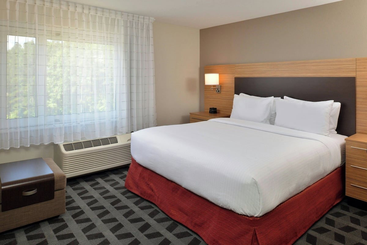 Une chambre d’hôtel avec un grand lit, une table de chevet, une commode, un radiateur et une fenêtre avec des rideaux légers.