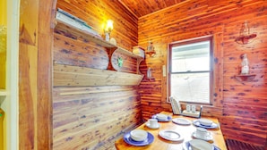 A wooden cabin interior with a dining table set for four, a window with blinds, and wooden walls. - Eureka Springs Vacation Rental < Half-Mi to Dtwn! (Eureka Springs)