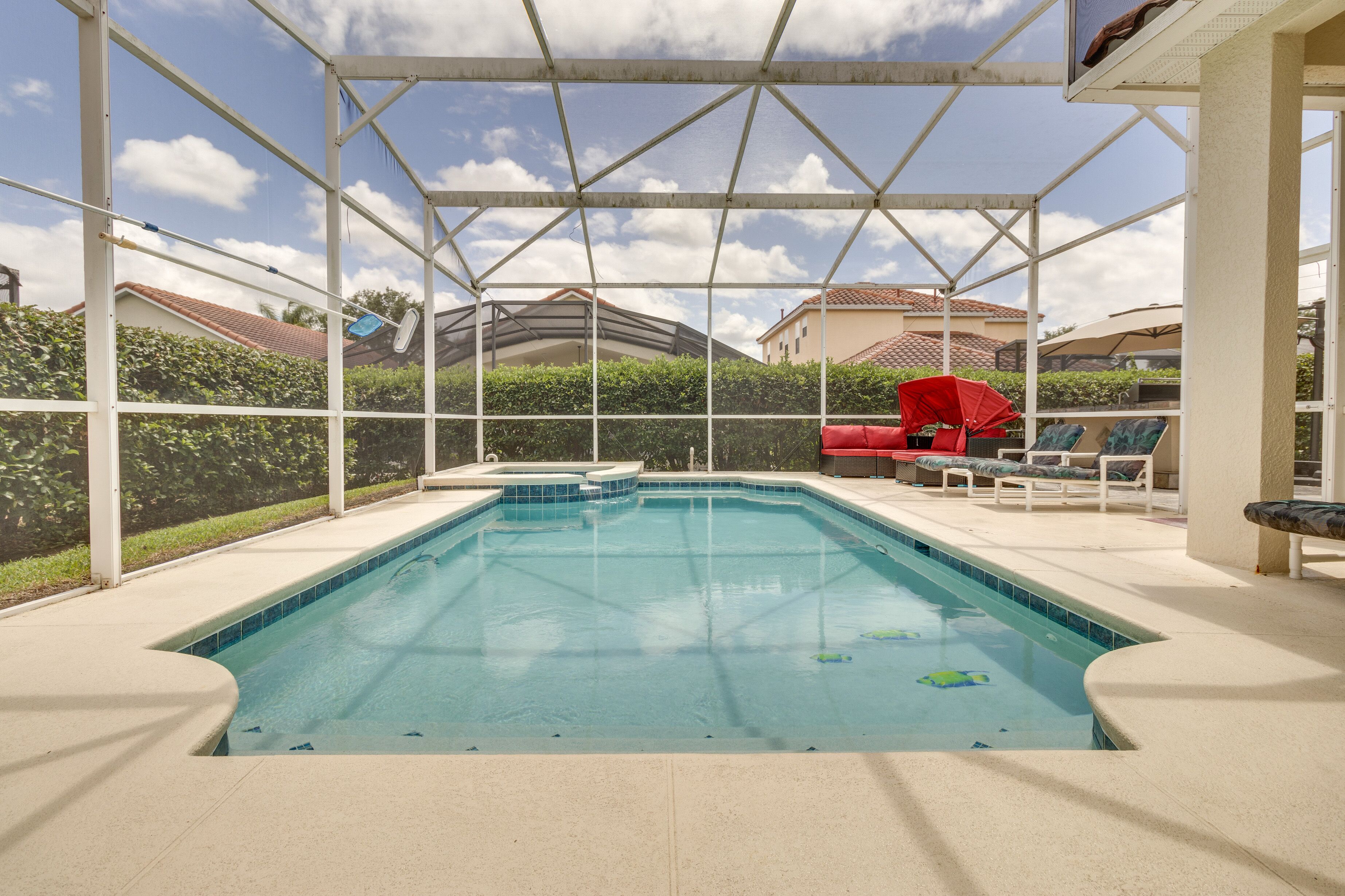 Ein überdachter Pool mit klar blauem Wasser, umrahmt von einem weißen Metallgestell und einer Fliesenbordsteinkante. Im Hintergrund befinden sich Liegestühle und ein roter Sonnenschirm.