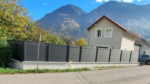 A single-story house with a red roof, a small front porch, and a garage. It is surrounded by a tall fence and set against a backdrop of mountains. - Escape and comfort 8-bed house with swimming pool (Val de Chaise)