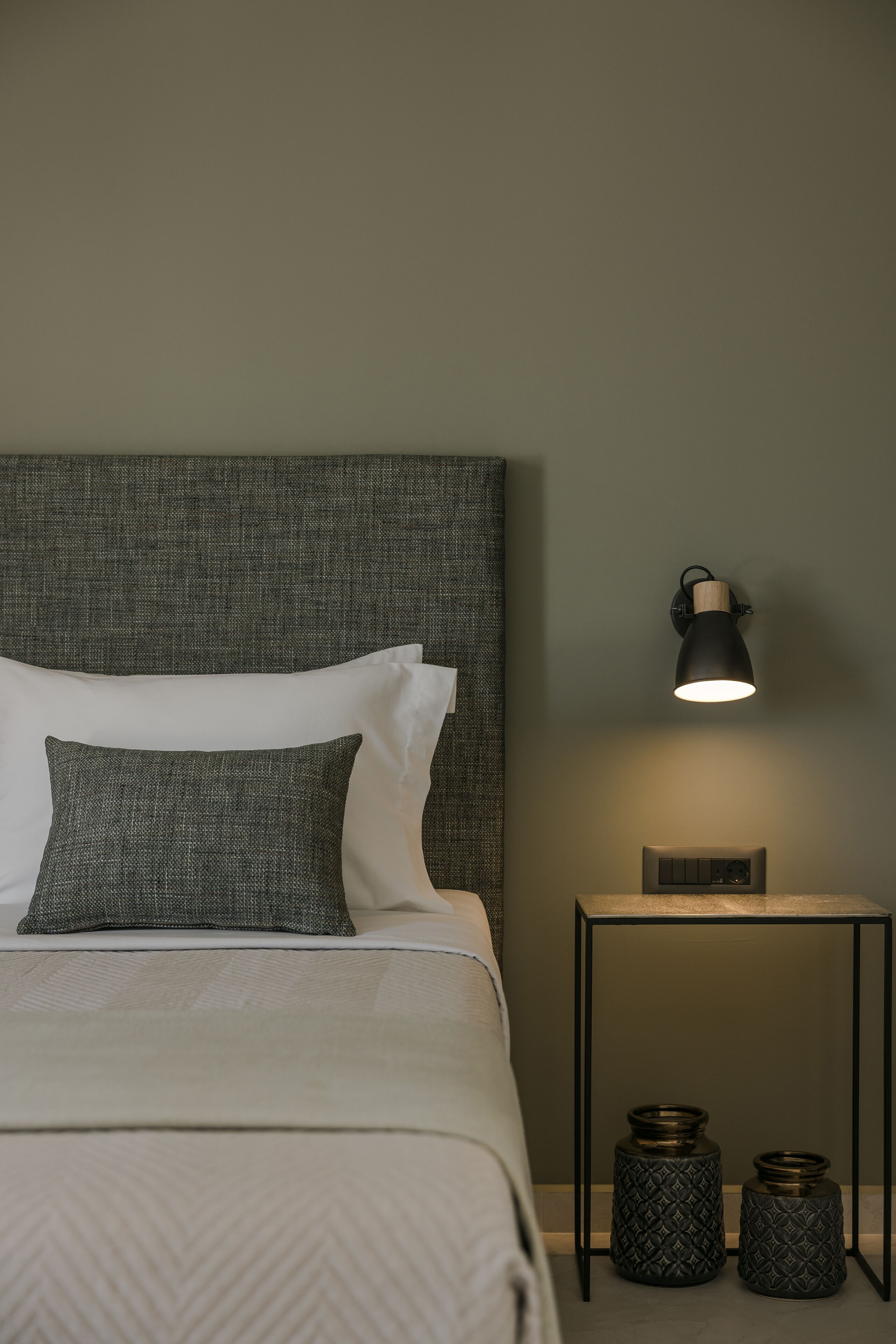 A neatly made bed with a grey headboard, a bedside lamp, and two decorative vases on a small table beside the bed.