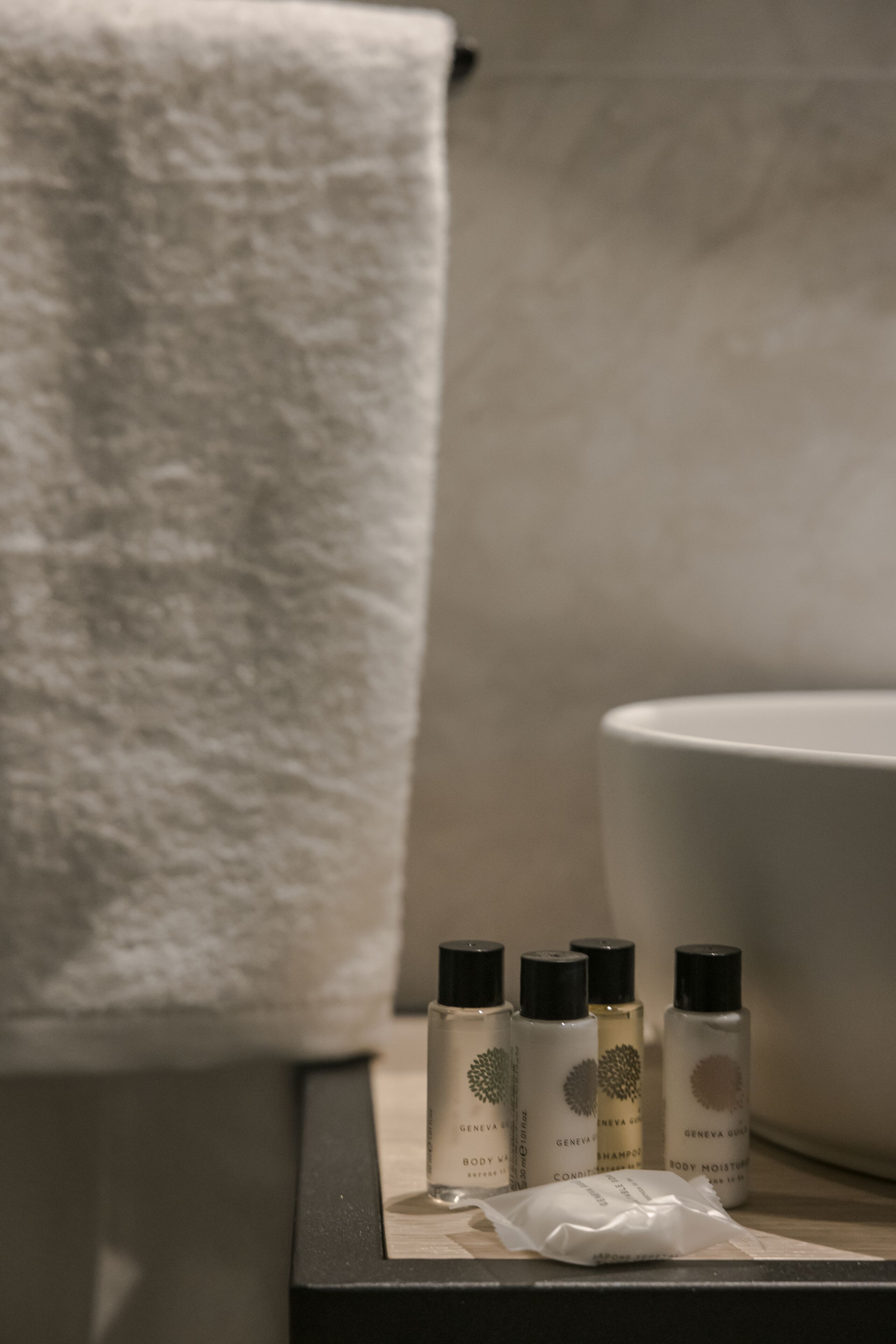Une salle de bain avec une serviette blanche suspendue à un porte-serviettes, un lavabo et trois flacons de produits de soins corporels disposés sur un plateau.