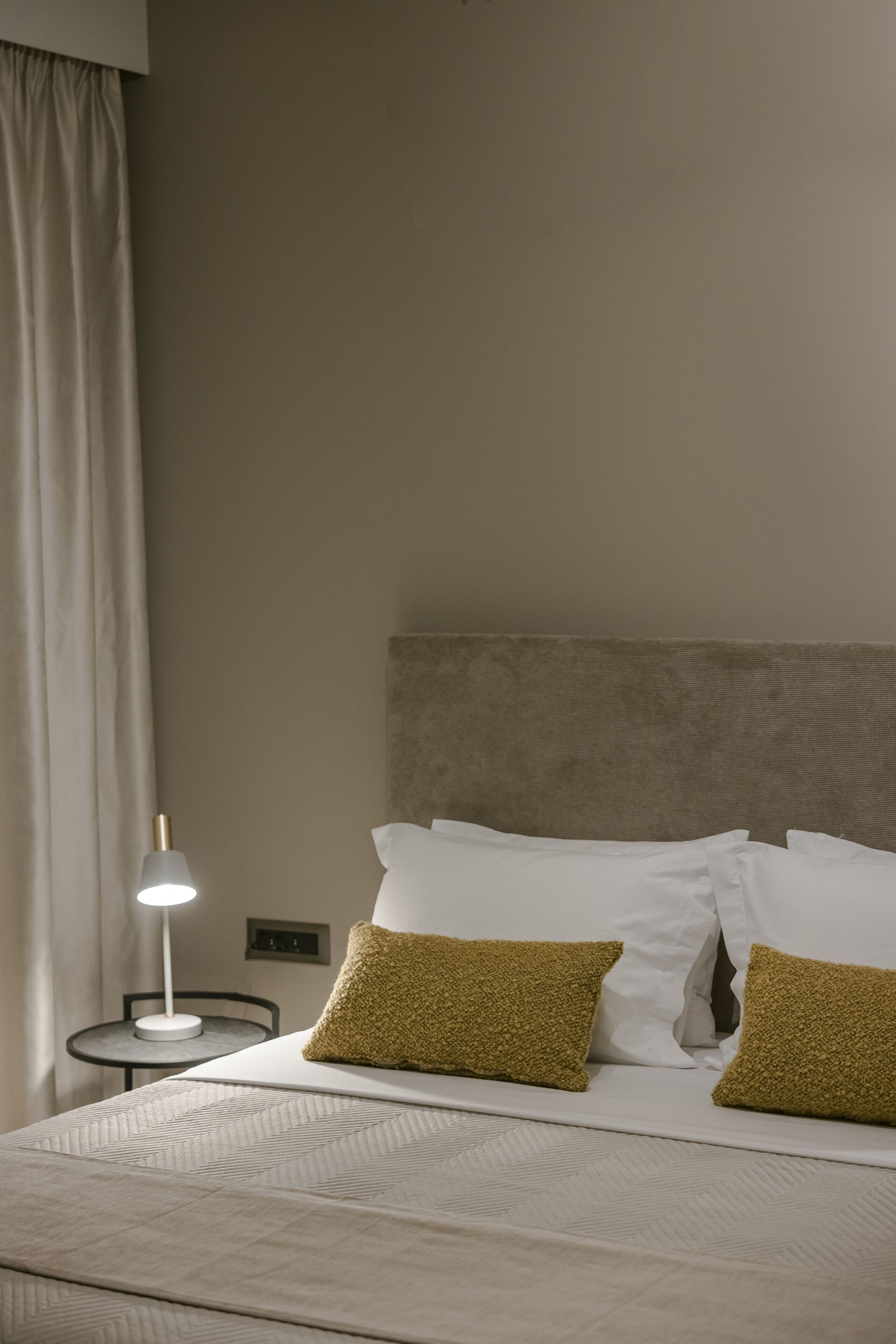 A neatly made bed with white linens and two textured pillows, a bedside lamp, and a small table with a lamp.