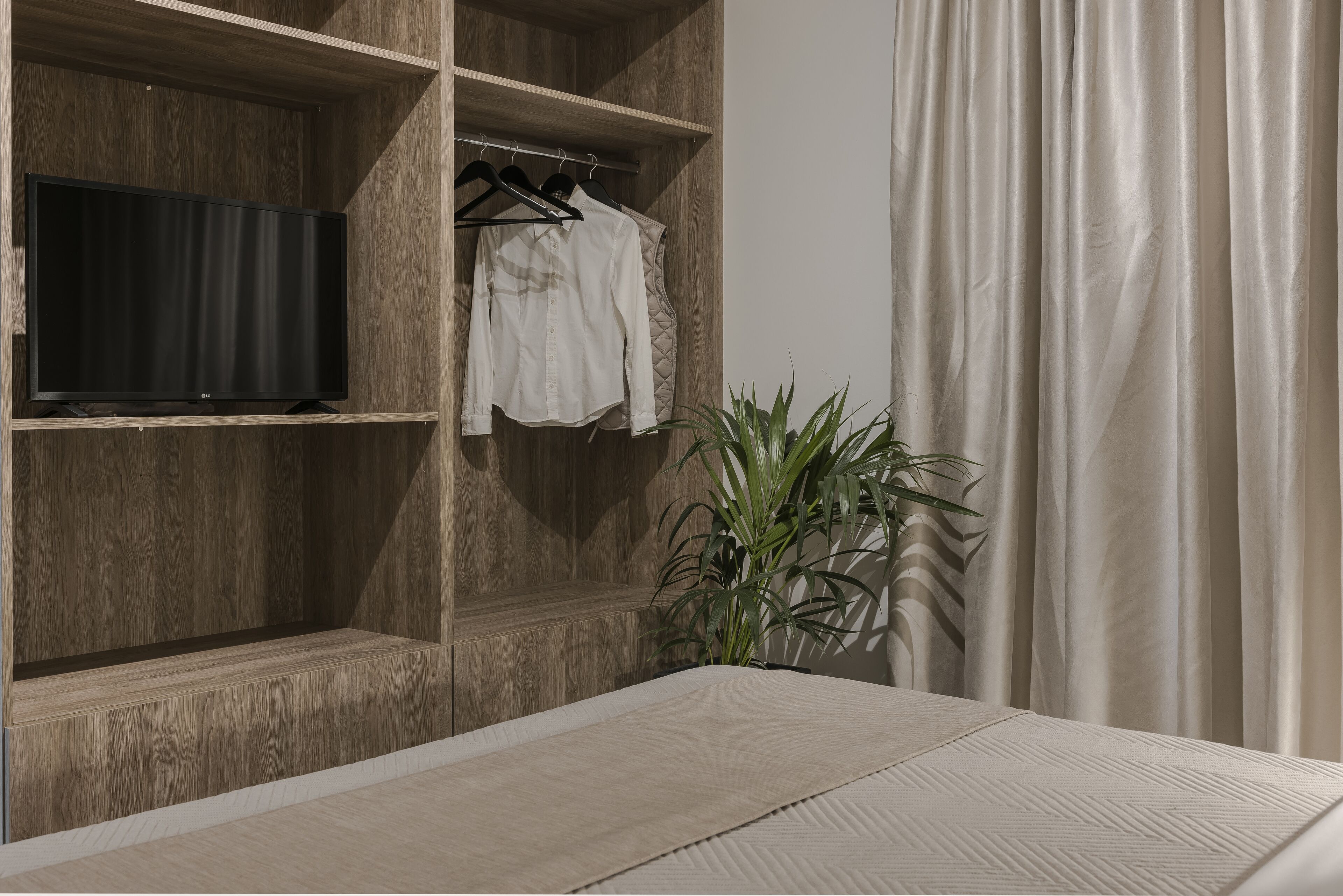 Un dormitorio con cama, televisión sobre una estantería de madera, perchero y planta en maceta.