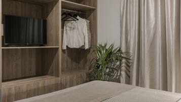 Un dormitorio con cama, televisión sobre una estantería de madera, perchero y planta en maceta.